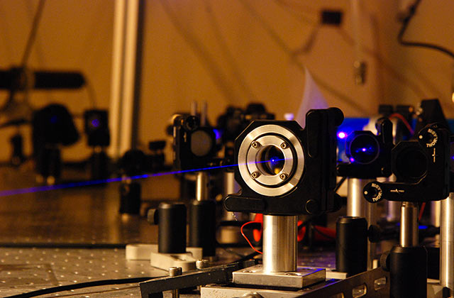 Équipement optique de laboratoire avec lentilles et miroirs montés sur banc optique, un faisceau laser bleu traverse la scène dans une ambiance tamisée.